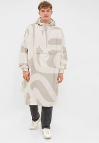 Ploaie în nuanțe de bej și gri, cu glugă, buzunar frontal și umeri căzuți, purtată peste blugi închiși la culoare și adidași albi.