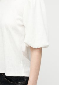 Blouse blanche à manches courtes bouffantes texturées portée avec un jean en denim noir, vue de profil, mettant en avant la manche et le bras.