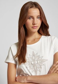 T-shirt bianco in cotone con un design centrale di una stella ricamata in beige metallico. Maniche corte e scollatura rotonda. Tessuto liscio.