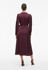 Woman with hair in a bun wearing a long-sleeve, belted, dark purple midi dress and dark high-heeled sandals, standing facing away.