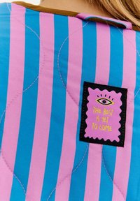 Kurtka pikowana z pionowymi niebieskimi i różowymi paskami, z brązowym kołnierzem i czarną metką z napisem "Najlepsze jeszcze przed nami."