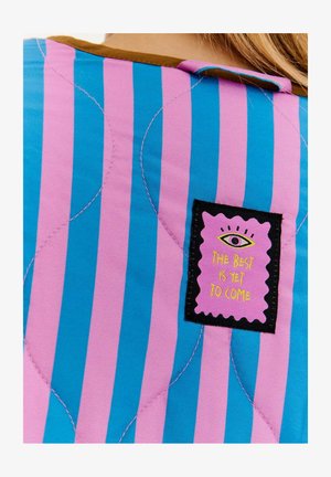 Prešívaný odev s vertikálnymi modro-ružovými pruhmi, s hnedým golierom a čiernym štítkom s nápisom "To najlepšie ešte len príde."