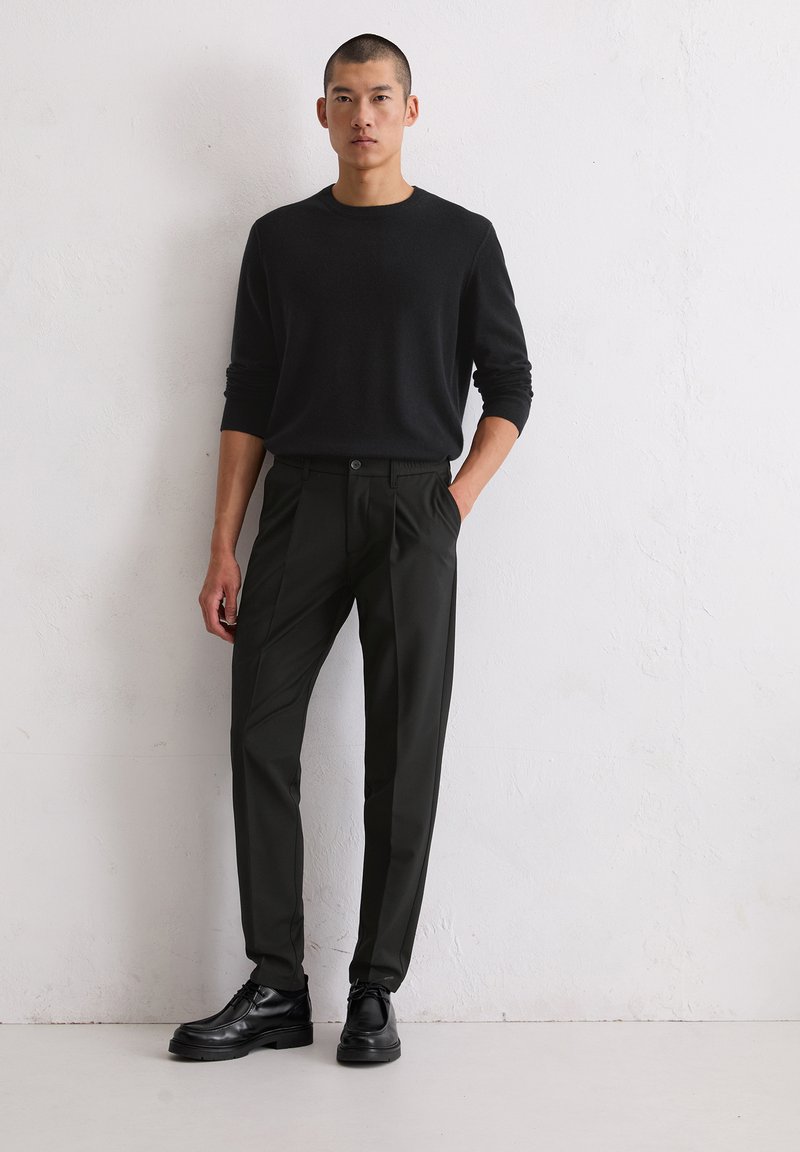 Zwarte blouse met lange mouwen gecombineerd met donkere groene op maat gemaakte broeken en zwarte schoenen. De outfit heeft een slim fit en strakke lijnen.
