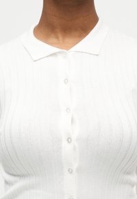 Κοντινό πλάνο ενός ατόμου που φοράει λευκό ριμπ πουκάμισο με κουμπιά και γιακά, εμφανίζοντας το άνω μέρος του στήθους και την περιοχή του λαιμού.