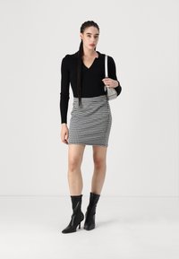 Fekete bordázott hosszúujjú felső, kutyafog mintás mini szoknya és elegáns fekete bokacsizma. Az ezüst kézitáska kontrasztot ad. Semleges háttér.