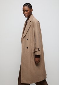 Beige wool blend double-breasted coat with notch lapels, two black buttons, and side pockets. Model wears a brown sweater and trousers.