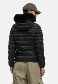 Giubbotto puffer nero con cappuccio foderato in pelliccia sintetica, design trapuntato e polsini elasticizzati. Abbinato a pantaloni cargo beige.