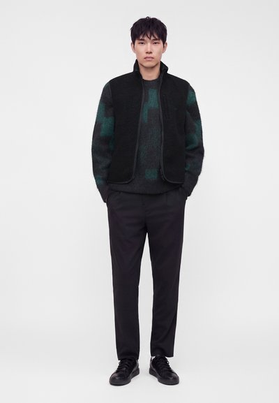 Άνδρας που φοράει μαύρο γιλέκο με φερμουάρ πάνω από ένα σκούρο πράσινο και μαύρο πουλόβερ με σχέδια, μαύρο παντελόνι και μαύρα αθλητικά παπούτσια, όρθιος με τα χέρια στις τσέπες.