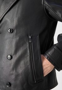 Musta nahkatakki, jossa on kaksirivinen malli, teksturoitu pinta, suuret napit ja vetoketjullinen sivutasku käytännöllisyyden lisäämiseksi.