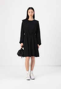 Black dress with lace trim, long sleeves, and an elastic waist. Model holds a black drawstring bag with floral accents and wears white sneakers.