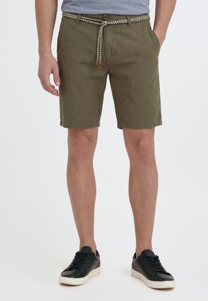 Homme portant un short vert olive avec une ceinture tissée, un t-shirt gris et des baskets noires avec semelles blanches sur fond blanc.