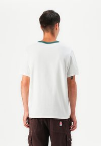Camiseta blanca de algodón con un cuello acanalado verde, mangas cortas y un corte relajado, combinada con pantalones de pana marrón oscuro.