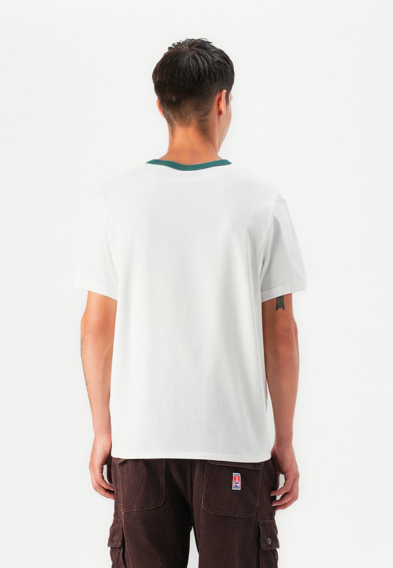 Biała bawełniana koszulka z zielonym ściągaczem przy dekolcie, krótki rękaw i luźny krój, noszona z ciemnobrązowymi spodniami z korduroju.