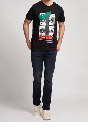 Camiseta de algodón negra con estampado gráfico en blanco, verde y rojo; combinada con pantalones vaqueros azul oscuro con detalles desgastados y zapatillas blancas.