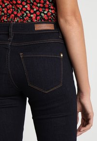 Mörkblå denimjeans med en åtsittande design, med en brun läderetikett på midjan och guldstickningar på fickorna.