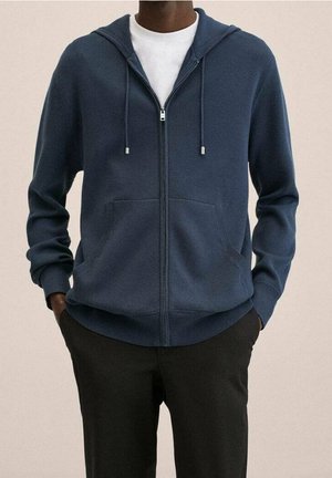 Homme portant un sweat à capuche bleu marine zippé par-dessus un t-shirt blanc, les mains dans les poches d’un pantalon noir, debout devant un fond uni.
