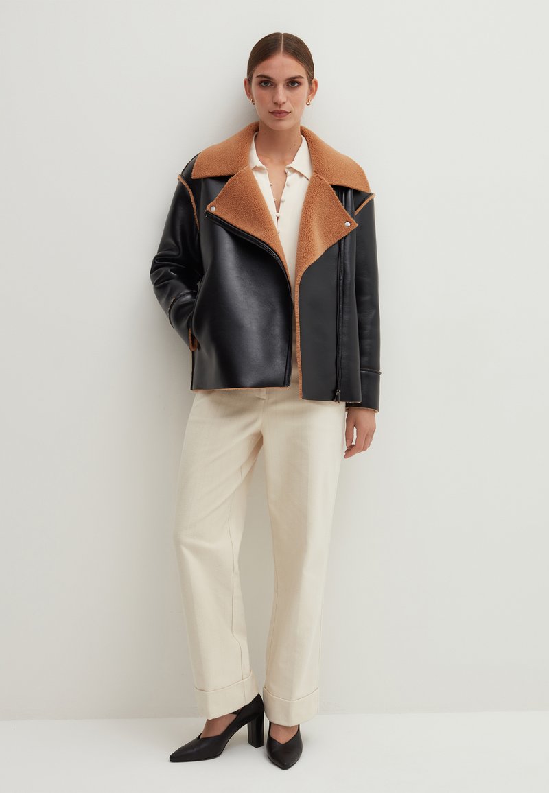 Schwarze Lederjacke mit einem hellbraunen Kunstfellkragen, Reißverschluss und seitlichen Taschen, kombiniert mit cremefarbenen Hosen und schwarzen High-Heels.