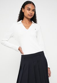 Λευκό πουκάμισο με μακριά μανίκια και V-λαιμόκοψη, που διαθέτει μικρό λογότυπο στο στήθος, σε συνδυασμό με σκούρα μπλε πλισέ φούστα.