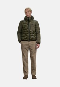 Pufferjack in donkergroen olijf met gestikte textuur, rits aan de voorkant, capuchon en borstzak. Gecombineerd met beige broek en bruine schoenen.