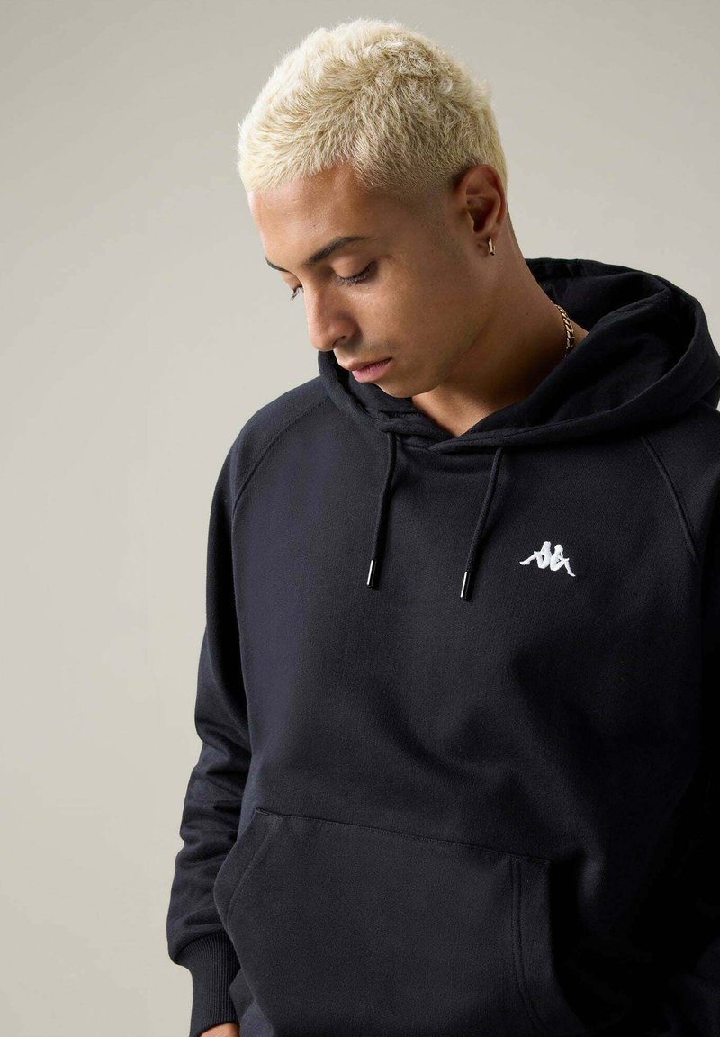 Jeune personne aux cheveux courts décolorés portant un sweat à capuche noir avec un logo blanc, regardant vers le bas sur un fond clair uni.