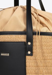 Sac en toile beige avec des anses en cuir noir et une poche, fermeture à cordon beige en tissu, et petit logo de marque doré.