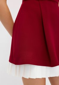 Robe courte avec un corsage bordeaux ajusté et une jupe plissée blanche superposée en bas. Texture lisse, sans motifs ni éléments métalliques visibles.