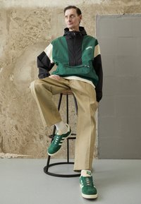 Windbreaker in green, black, and cream; tan wide-leg pants; green suede sneakers with cream accents; sits on a black metal stool.