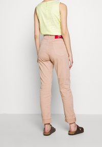 Vaalean keltainen hihaton paita, jossa on takana napit, yhdistettynä käärittyihin vaaleanpuniin housuihin. Ruskeat sandaalit, joissa on rakenteellinen pohja.