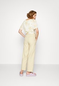 Blusa floral em creme com mangas curtas, combinada com calças bege de cintura alta e sandálias lilás claras. Vista traseira.