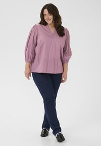 Smilende kvinde med langt brunt hår iført en pink bluse med 3/4 puffede ærmer, mørkeblå jeans og sorte loafers på en hvid baggrund.