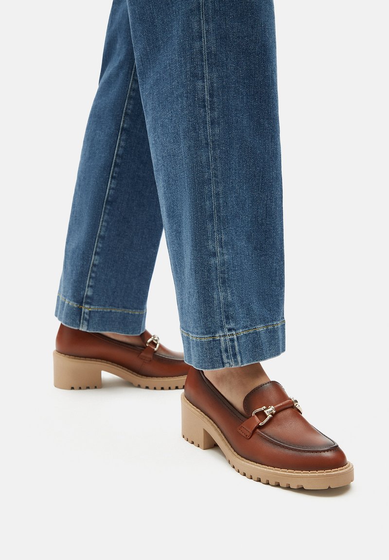 Hnědé kožené mokasíny s kulatou špičkou, dekorativním páskem, stříbrným kováním a béžovou hrubou podrážkou. V kombinaci s džínovými kalhotami.