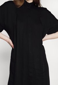 Robe noire à boutons, avec des manches courtes en dolman, texture lisse, plis sur les côtés et col haut. Design simple et ample.