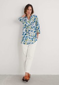 Een button-up overhemd met een blauw bloempatroon, voorzien van gele en groene accenten. Draag het met witte broeken en bruine klompen.