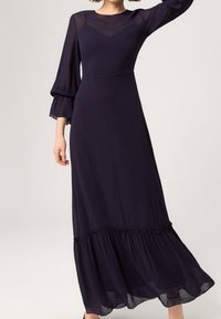 Robe longue navy avec des manches superposées transparentes, un corsage ajusté et un ourlet à volants. Texture lisse avec un léger éclat.