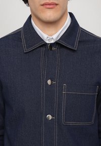 Homme portant une veste en jean bleu foncé avec des boutons-pression argentés et une chemise blanche à col en dessous.