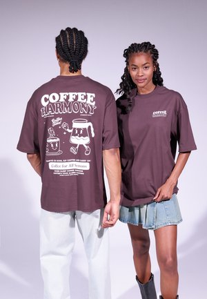 Le t-shirt oversized in cotone marrone presentano grafiche "COFFEE HARMONY" sul retro, con illustrazioni giocose a tema caffè e accenti di testo bianchi.