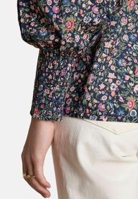 Sievietes roka ar zelta gredzeniem, tērpta tumši zila ziedu garo piedurkņu blūzē ar smocka aproci un off-white biksēm.