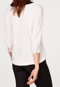 Hvid blouse med trekvartærmer og sort krave. Let, glat stof med knapdetalje ved ærmeafslutningerne. Enkel, ren design.