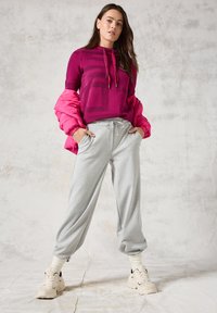 Rosa Kapuzenpullover mit geometrischem Design, kombiniert mit grauen Jogginghosen. Das Modell trägt beigefarbene Sneakers. Weiche Materialien und lässige Passform.