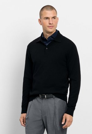 Mann mit kurzen Haaren, der ein schwarzes langärmeliges Polohemd über einem dunkelblauen Hemd mit Kragen und grauen Hosen mit schwarzem Gürtel trägt.