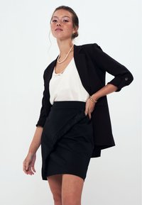 Gonna nera con orlo frangiato, abbinata a una camicia bianca e a un blazer nero. La modella posa con una collana di perle e un bracciale.