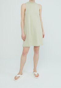 En ljus olivgrön ärmlös klänning med hög halsringning, som har en avslappnad passform och en mjuk, slät textur. Vita flip-flops fulländar looken.