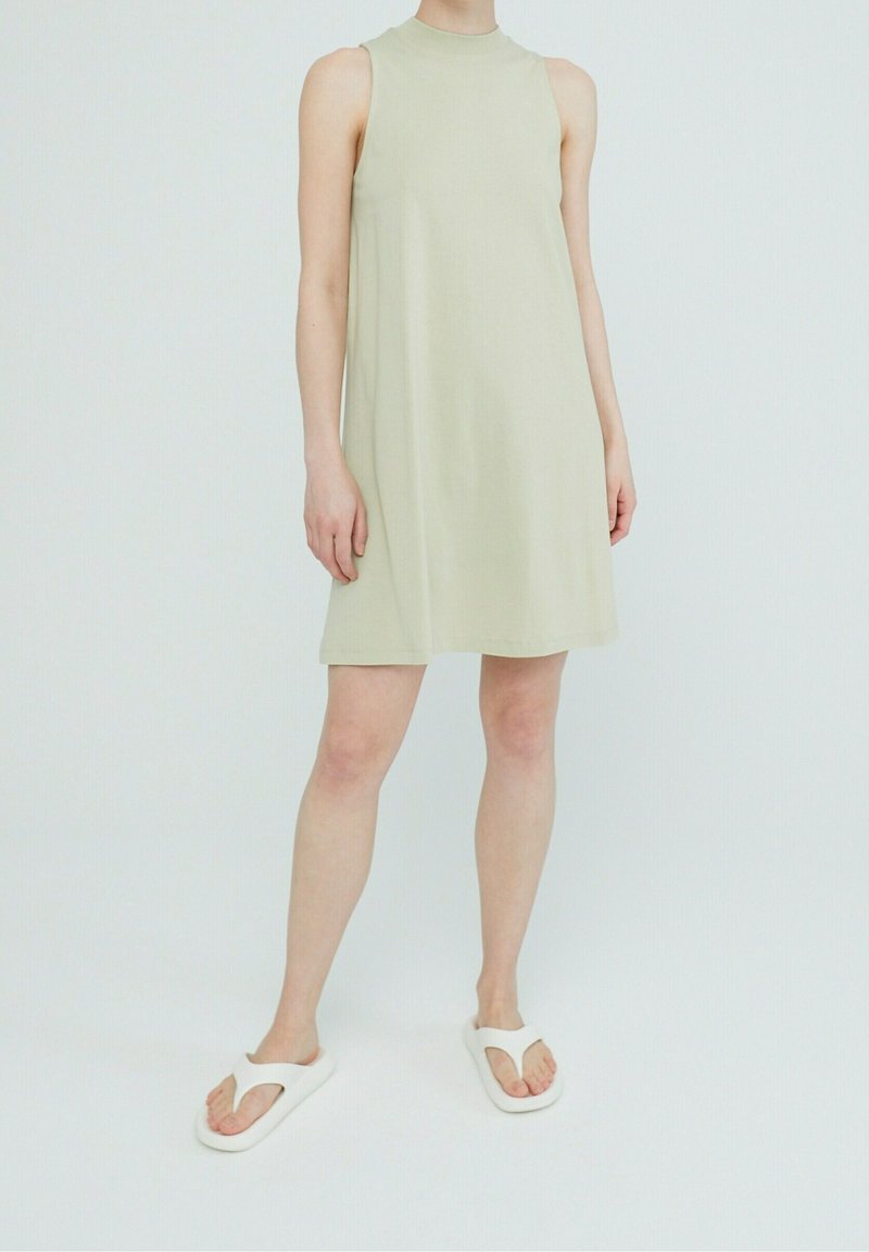 En ljus olivgrön ärmlös klänning med hög halsringning, som har en avslappnad passform och en mjuk, slät textur. Vita flip-flops fulländar looken.