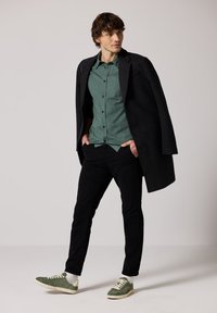 Černý vlněný kabát přes zelenou kostkovanou košili, doplněný o černé kalhoty a zelené semišové tenisky. Slim fit a ležérní design.