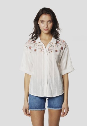 Camisa blanca a rayas con mangas cortas, con bordado floral en rosa, amarillo y verde en los hombros y cierre frontal con botones.