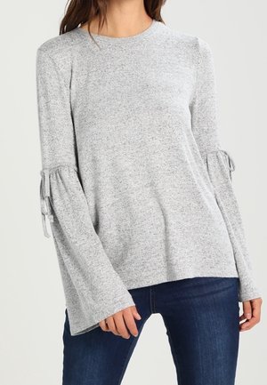 Kobieta w jasnoszarym swetrze z długimi rozkloszowanymi rękawami i detalami z wiązaniami, w połączeniu z ciemnoniebieskimi jeansami.