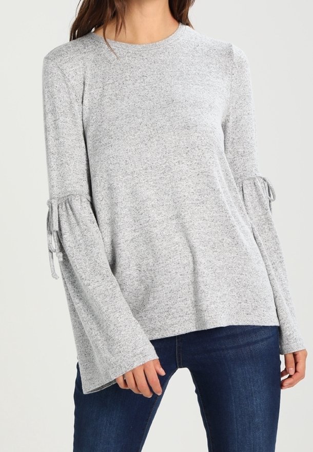 Femme portant un pull gris clair à manches longues évasées avec des détails à nouer, associé à un jean bleu foncé.