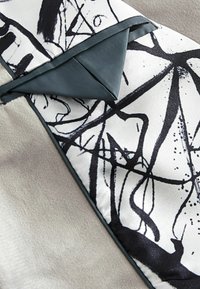 Tejido texturizado de base gris; presenta un patrón abstracto en blanco y negro. Se observa un acento doblado en un material azul-verde a juego.
