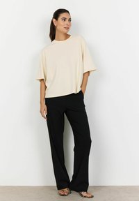 Bežové oversized tričko s okrúhlym výstrihom, spárované s čiernymi nohavicami. Model má na sebe vzorované sandále a stojí na jednoduchom pozadí.