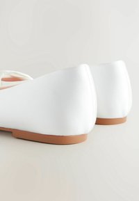 Ballerines en satin blanc avec une finition lisse, un dos arrondi et une semelle en caoutchouc marron. Présentent des coutures minimales et un design épuré.